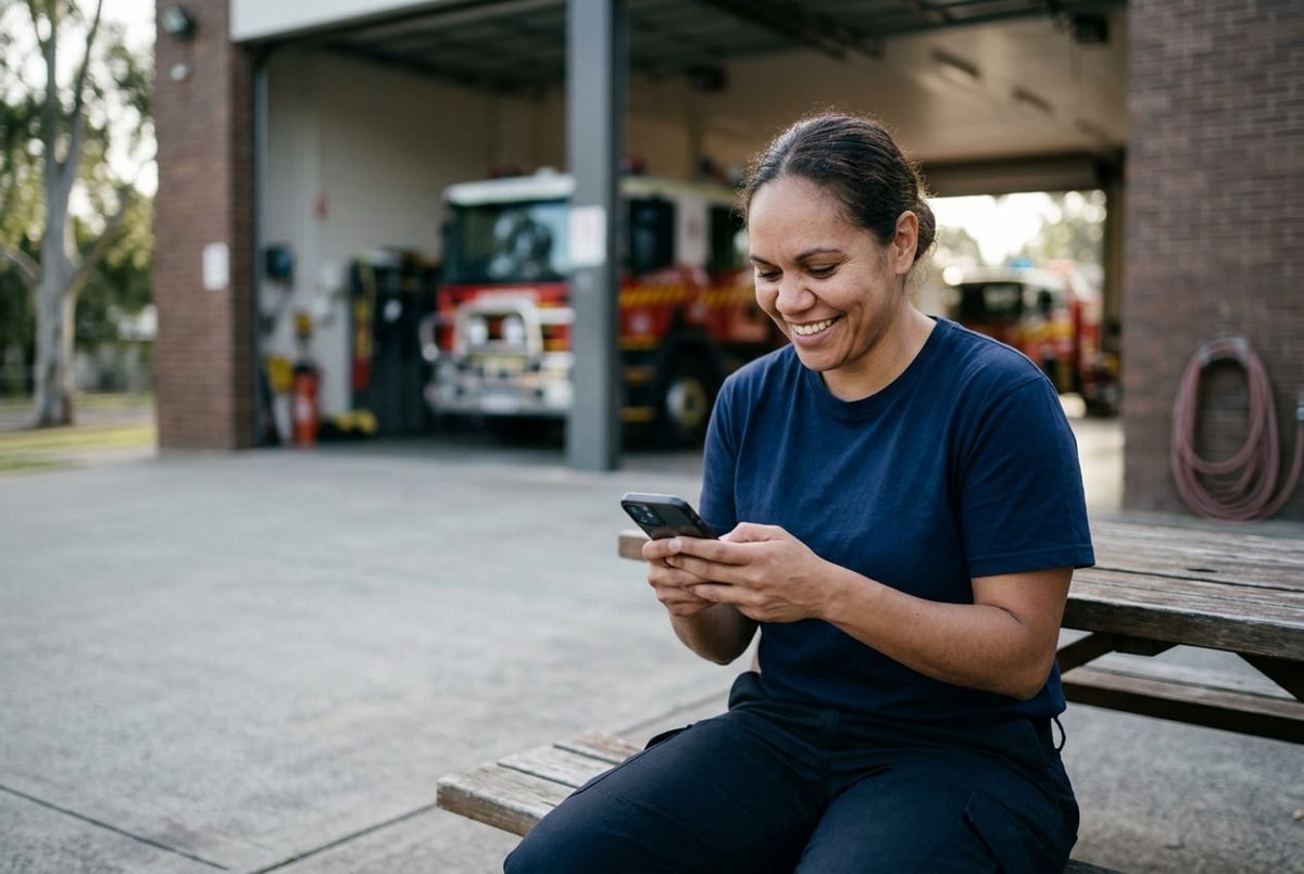 Woman in a navy t-shirt smiling at her phone on a bench outside a fire station